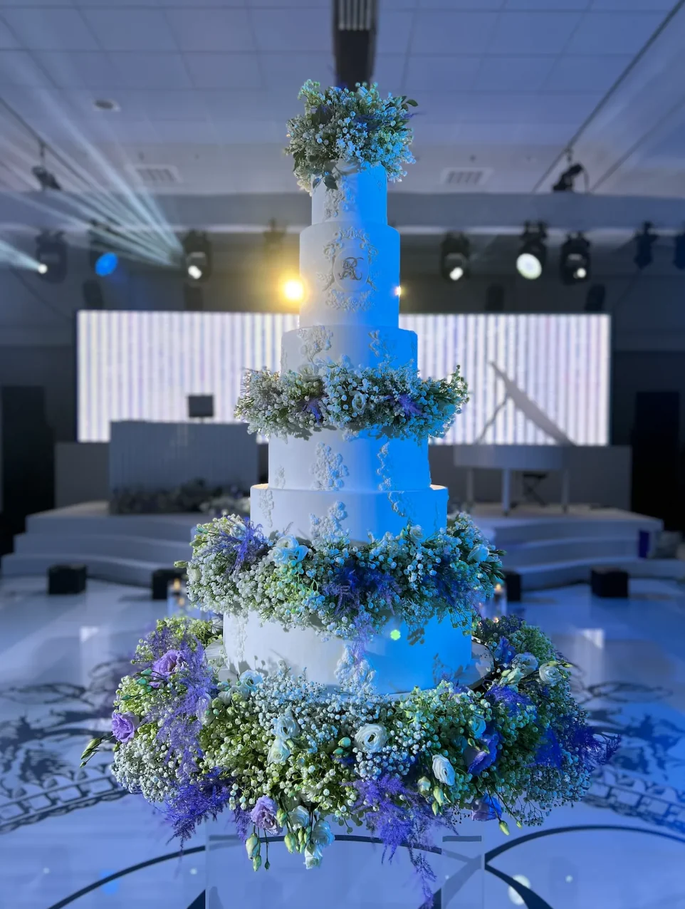 Enchanted Garden tall white wedding cake encircled by flowers and greenery under blue reception lighting.