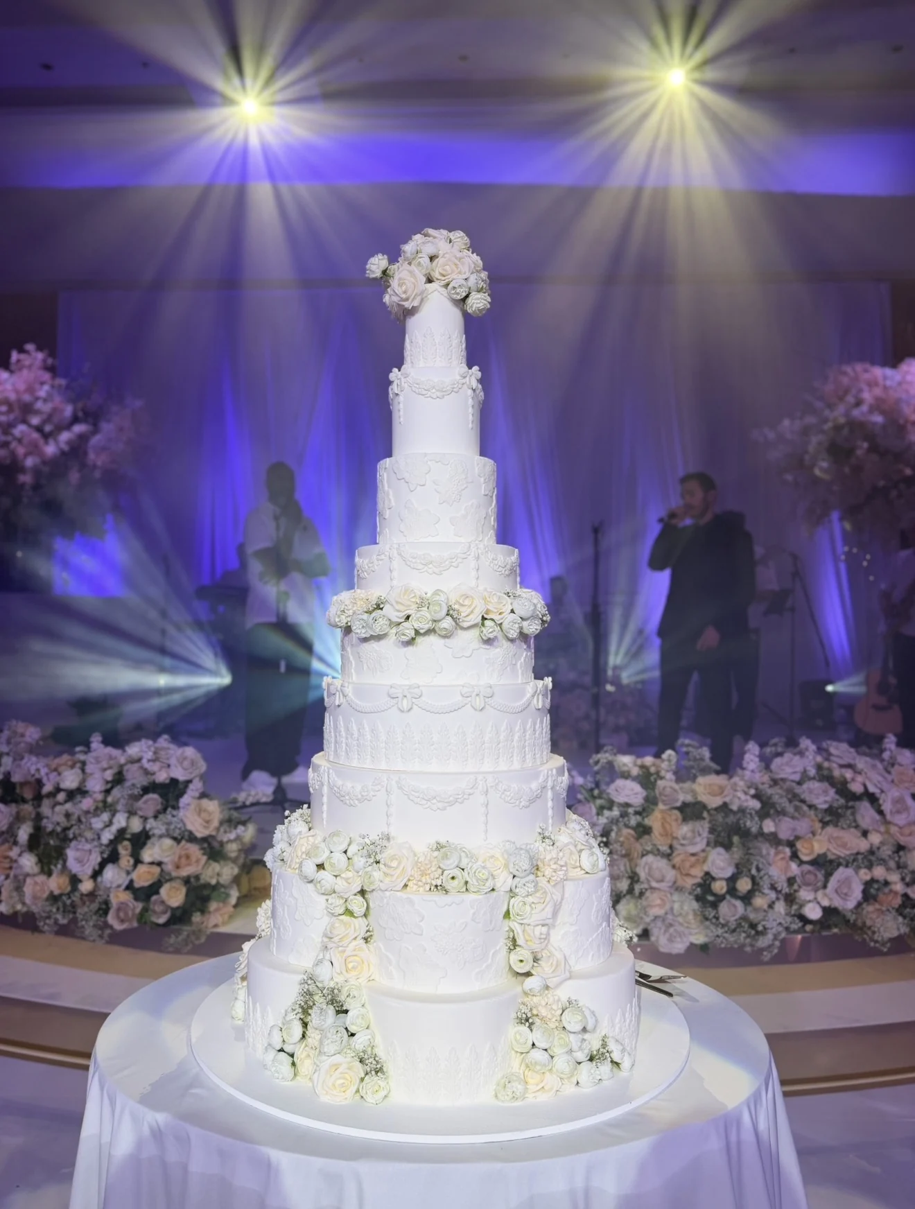 Ophelia tall white wedding cake with floral cascades and decorative tiers beneath golden spotlights.