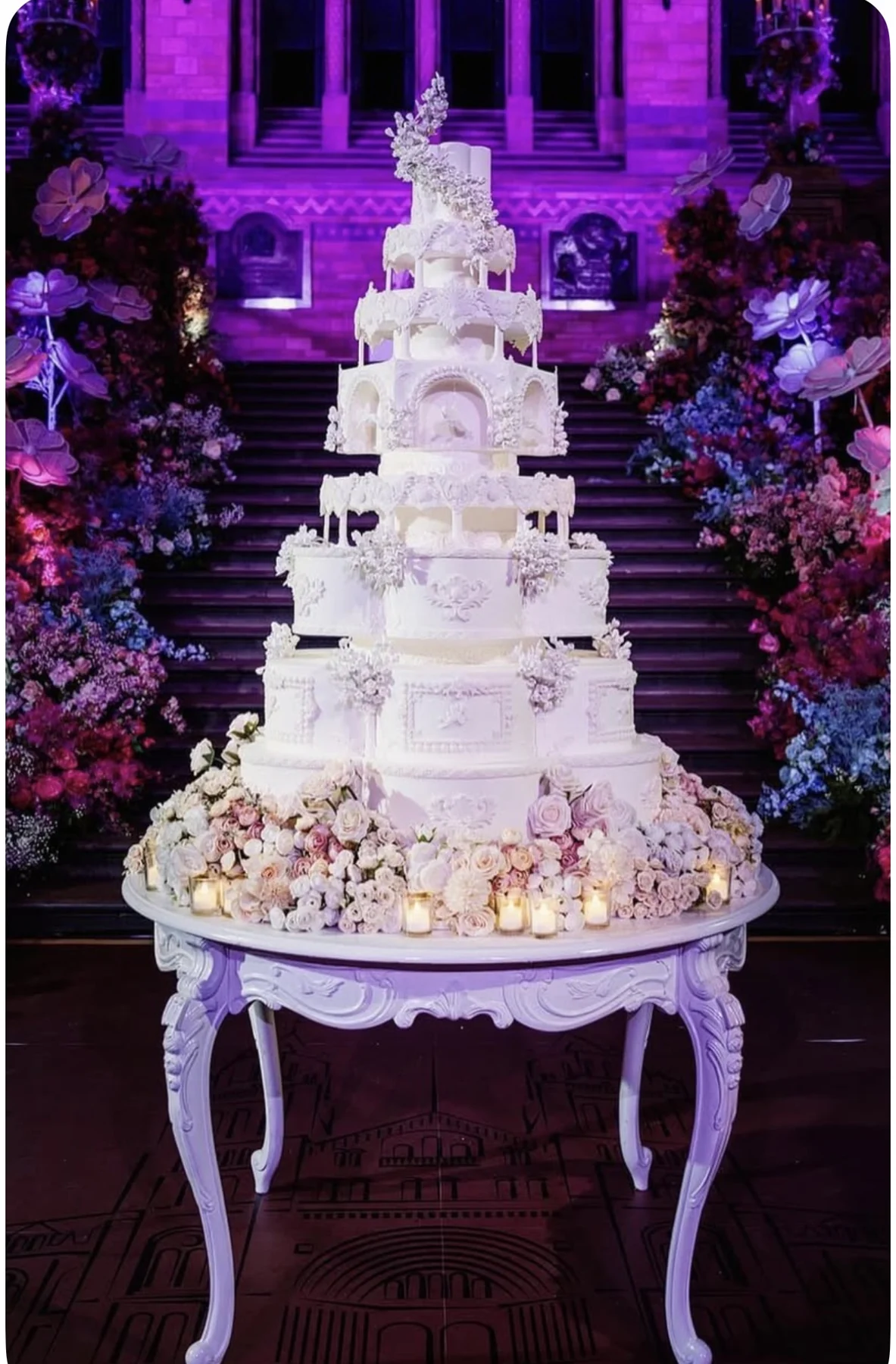 Venetian ornate ivory wedding cake with arches and floral details on a carved table before a grand staircase.
